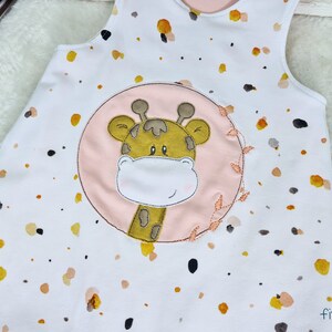May include: White sleeveless onesie with a pink circle and a brown giraffe applique. The onesie has a brown, yellow, and orange splatter pattern.