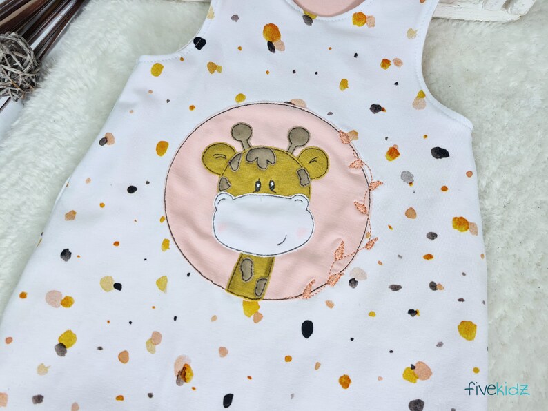 May include: White sleeveless onesie with a pink circle and a brown giraffe applique. The onesie has a brown, yellow, and orange splatter pattern.