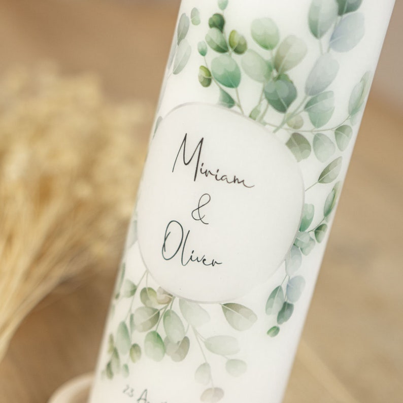 May include: White candle with a watercolor design of green eucalyptus leaves. The candle has a white oval label with the text "Miriam & Oliver" in black calligraphy.
