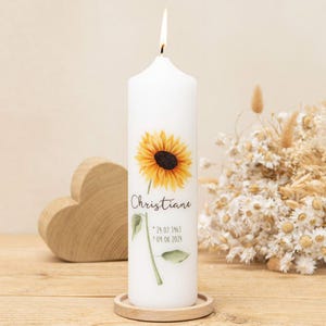 Op de afbeelding: Een witte kaars met een brandende lont, met een zonnebloemontwerp en de naam "Christiane" met data. De kaars staat op een houten onderzetter. Een houten hart en droogbloemen staan op de achtergrond.