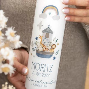 Könnte beinhalten: Eine weiße Kerze mit einer farbenfrohen Illustration der Arche Noah mit Tieren, einem Regenbogen und dem Namen "MORITZ" sowie dem Datum "5. JULI 2022". Die Kerze enthält auch einen Bibelvers. Ein dekorativer Artikel für eine Taufe oder Konfirmation.