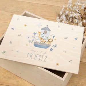 May include: A wooden box with a light wood lid. The lid has a blue and white illustration of a boat with animals on it. The text "Meine Taufe MORITZ" is printed on the lid.