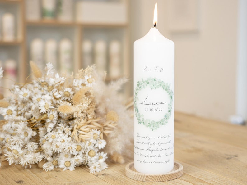 May include: White baptism candle with a green eucalyptus wreath design. The candle has the name "Luca" and the date "26.10.2022" printed on it. The text "Zur Taufe" is also printed on the candle.