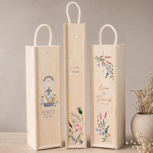 May include: Three tall, rectangular light-colored wooden gift boxes with white rope handles. The left box features a Noah's Ark illustration with the text MORITZ 5. JULI 2022. The center box shows floral art and the text Lara 16. Mai 2026. The right box displays floral designs and the text Lena & Daniel 14.08.2023.