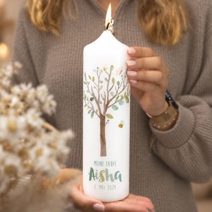 Op de afbeelding: Een witte stompkaars met een brandende vlam, voorzien van een aquarelillustratie van een boom met groene bladeren, een kleine vlinder en een bij. De tekst MEINE TAUFE, Aisha en 8. MAI 2024 staat in groene en grijze letters op de voorzijde.