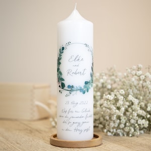 Könnte beinhalten: Weiße Hochzeitskerze mit grünem floralem Kranzdesign. Die Kerze trägt die Namen "Elke und Robert" und das Datum "28. August 2022". Der Text lautet "Was far in Glack von du jemanden findest der so ganz gern in dein Herz passt."