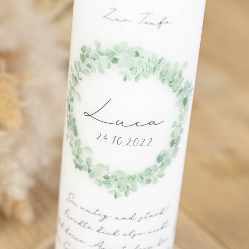 May include: White candle with a green eucalyptus wreath design. The text "Zur Taufe" is at the top, and the name "Luca" and the date "24.10.2022" are in the center. The text "Sei mutig und stark!" is at the bottom.