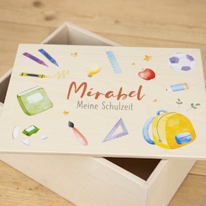 Può includere: Una scatola di legno con una finitura in legno chiaro. Il coperchio presenta un design colorato con materiale scolastico, un pallone da calcio, una mela e uno zaino. Il testo sul coperchio recita "Mirabel Meine Schulzeit".