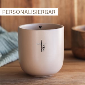 Könnte beinhalten: Eine beige Keramiktasse mit einem schwarzen Kreuz und dem Wort "HOLY" vertikal geschrieben. Ein kleines schwarzes Herz ist oben an der Tasse zu sehen.