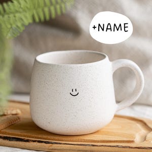 Könnte beinhalten: Eine gesprenkelte, weiße Keramiktasse mit runder Form und großem Henkel. Auf der Vorderseite befindet sich ein schwarzes Smiley-Gesicht. Der Text "+NAME" steht in einer weißen Sprechblase über der Tasse.