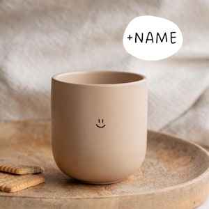 Könnte beinhalten: Eine beige Keramiktasse mit rundem Boden und einem einfachen Smiley. Die Tasse steht auf einem Holztablett mit zwei Keksen. Ein weißes Oval mit dem Text "+NAME" befindet sich über der Tasse.