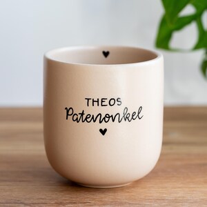 Könnte beinhalten: Beige Keramik-Pflanztopf mit dem Text "THEOS Patenonkel" in schwarzer Schrift und zwei kleinen schwarzen Herzsymbolen. Der zylindrische Topf steht auf einer Holzoberfläche.