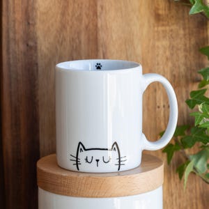 Könnte beinhalten: Weiße Keramiktasse mit einer schwarzen Katzenkopf-Illustration. Die Katze hat geschlossene Augen und ein kleiner Pfotenabdruck ist auf der Oberseite der Tasse sichtbar.