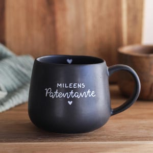 Könnte beinhalten: Ein mattschwarzer Keramikbecher mit abgerundeter Form und einem Henkel. Auf der Vorderseite befindet sich der weiße Schriftzug "MILEENS Patentante", eingerahmt von zwei kleinen weißen Herzsymbolen. Ein kleines weißes Herz ist am Innenrand zu sehen.