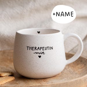 Könnte beinhalten: Eine gesprenkelte, weiße Keramik-Tasse mit runder Form und großem Henkel. Auf der Tasse steht in schwarzer Schrift "THERAPEUTIN mit", mit einem Herzsymbol darüber und darunter. Ein weißer Kreis mit "+NAME" befindet sich darüber.