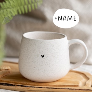 Könnte beinhalten: Eine gesprenkelte weiße Keramiktasse mit einem schwarzen Herzdesign. Die Tasse hat eine abgerundete Form und einen großen Henkel. Der Text "+NAME" befindet sich in einem weißen Oval über der Tasse. Die Tasse steht auf einem Holzschneidebrett.