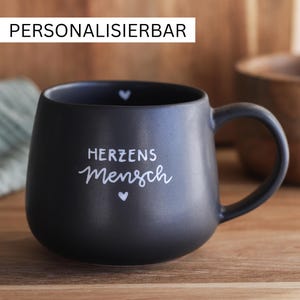 Könnte beinhalten: Eine mattschwarze Keramiktasse mit dem weißen Schriftzug "HERZENS mensch" und kleinen weißen Herzsymbolen auf der Vorderseite und am inneren Rand. Das Wort "PERSONALISIERBAR" steht in einem weißen Kasten über der Tasse.