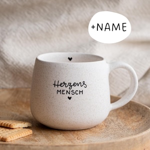 Könnte beinhalten: Eine gesprenkelte weiße Keramiktasse mit runder Form und Henkel. Auf der Tasse stehen die Worte "Herzens MENSCH" und ein Herzsymbol. Ein Holztablett und Kekse sind im Vordergrund.