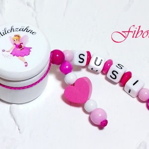 Può includere: Una scatola bianca con un'illustrazione di fata rosa e il testo "Milchzähne" sul coperchio. Un ciondolo a forma di cuore rosa e una collana di perline rosa e bianche con il nome "SUSI" scritto in lettere bianche.