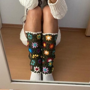 May include: A pair of green crochet leg warmers with colorful flower designs. The leg warmers are worn over white socks. The flowers are in shades of yellow, orange, pink, and blue. The leg warmers are a unique and stylish accessory.