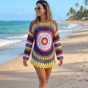 May include: A colorful, handmade crochet tunic dress with a circular design in the center. The dress features a rainbow of colors, including red, orange, yellow, green, blue, and purple. The long-sleeved dress is worn on a beach.