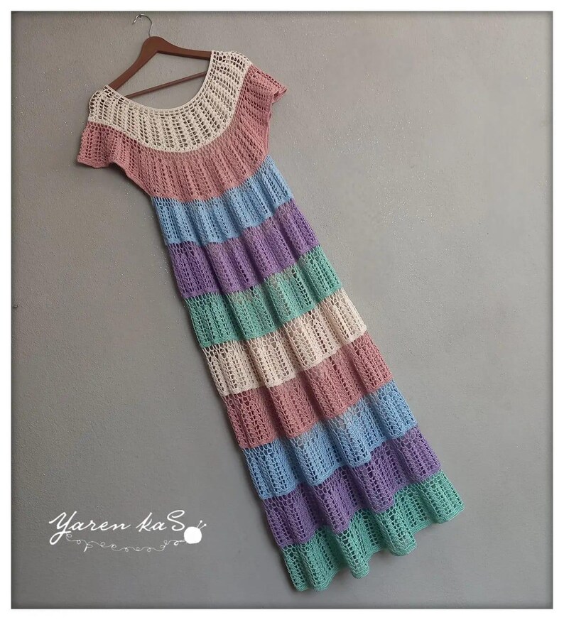 Pode incluir: Vestido longo de croch&ecirc; feito &agrave; m&atilde;o em cores pastel, incluindo rosa, azul, roxo e verde. O vestido tem um decote redondo e mangas curtas. O vestido est&aacute; pendurado em um cabide de madeira.