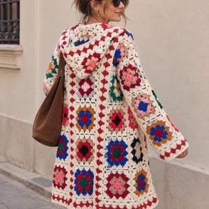 May include: A long-sleeved hooded cardigan made of cream-colored yarn featuring a multi-colored granny square crochet pattern. The squares include vibrant shades of red, blue, pink, green, and black, joined by a cream border throughout the garment.