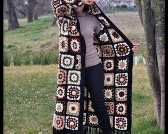 May include: A long, open-front crochet coat featuring a patchwork design of squares in cream, brown, burgundy, and black. The coat has a fringe trim along the bottom and is worn outdoors.
