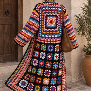 Op de afbeelding: Een lang, aan de voorzijde open vest gemaakt van veelkleurige gehaakte granny squares. Het kledingstuk heeft een groot vierkant patroon op de rug en kleinere bloemachtige vierkantjes aan de onderkant, alles omrand met zwart garen en een bruine tas.