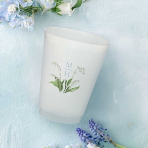 Op de afbeelding: Witte plastic beker met een bloemenontwerp en de initialen "M + H" gedrukt op de voorkant. De beker is omringd door blauwe en witte bloemen.