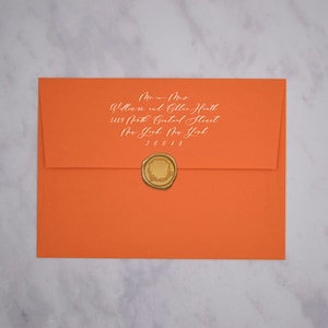May include: An orange envelope with a white address label that reads "Mr. & Mrs. William and Gillian Heath, 3189 North Central Street, New York, New York, 20018". A gold wax seal with a wreath design is on the envelope.