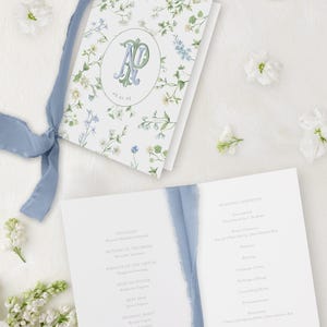 May include: A wedding ceremony program with a blue ribbon. The front cover features a floral pattern with a monogram in a circle. The inside pages list the officiant, mother of the bride, parents of the groom, maid of honor, best man, and wedding party.