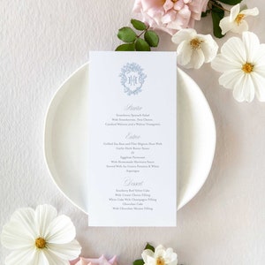 May include: A wedding menu card featuring a blue floral design and the initials "J.M.E." within a wreath. The menu lists starter, entree and dessert choices.