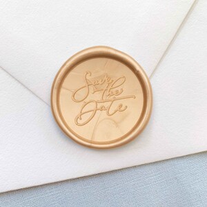 Puede incluir: Un sello de cera dorado con el texto "Save the Date" en escritura cursiva.