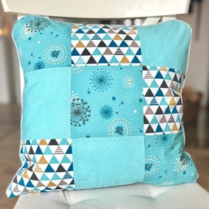 Könnte beinhalten: Ein türkisblaues Kissen mit Patchwork-Muster. Das Kissen besteht aus verschiedenen Stoffen mit geometrischen Mustern und einem Löwenzahndruck.