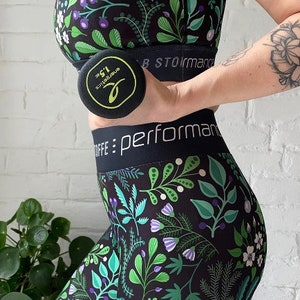 Könnte beinhalten: Eine Frau trägt schwarze Leggings mit einem Blumenmuster und einen schwarzen Sport-BH. Sie hält eine 1,5 kg schwere Hantel in ihrer rechten Hand. Die Leggings haben einen schwarzen Bund mit dem Text "caffe: performan"