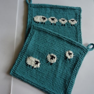 Könnte beinhalten: Zwei tealblaue gehäkelte Topflappen mit weißen Schafmotiven. Der größere Topflappen hat fünf Schafe in einer Reihe, während der kleinere drei Schafe hat.