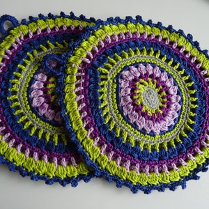 Könnte beinhalten: Zwei gehäkelte Topflappen in runder Form mit einem mehrfarbigen Design. Die Topflappen sind aus grünem, lila, blauem und grauem Garn gefertigt.