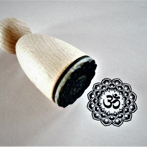 Könnte beinhalten: Holzstempel mit einem schwarzen Gummidesign eines Om-Symbols in einem Mandala. Der Stempel ist aus hellem Holz und hat eine abgerundete Oberseite.
