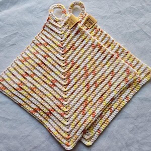 Könnte beinhalten: Zwei handgefertigte, quadratische Topflappen aus Häkelarbeit. Die Topflappen weisen ein Zickzackmuster in Weiß, Gelb, Orange und Braun auf. Jeder Topflappen hat eine Schlaufe zum Aufhängen und ein kleines Etikett mit der Aufschrift "Made with Love".