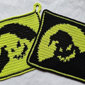 Könnte beinhalten: Zwei quadratische Topflappen aus Häkelarbeit. Einer ist schwarz mit einem neongrünen Design, der andere ist neongrün mit einem schwarzen Design. Jeder Topflappen hat ein lächelndes Gesichtsdesign und eine kleine Schlaufe zum Aufhängen.