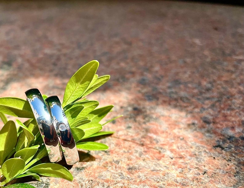 May include: Two silver wedding bands with a small diamond on one of the rings, resting on green leaves.