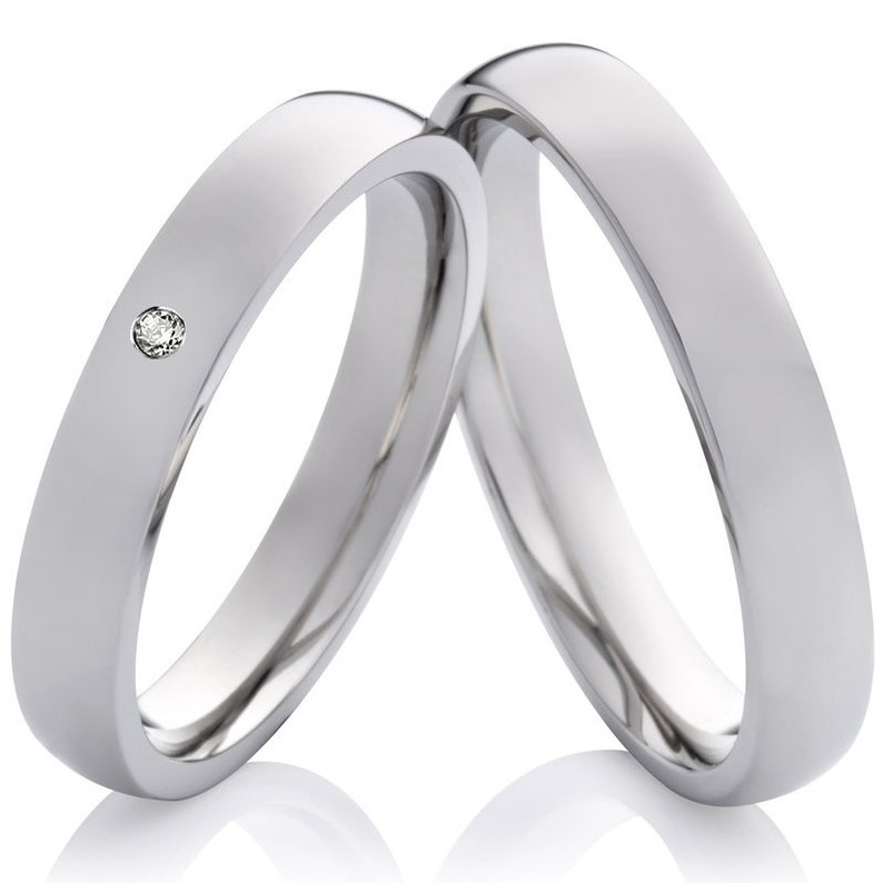 May include: Two silver wedding bands, one with a small diamond, are intertwined. The bands are smooth and polished.