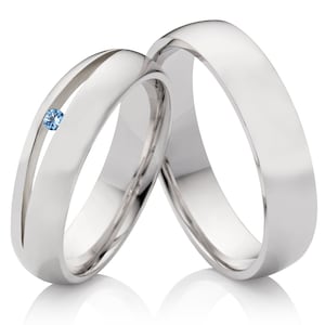 Op de afbeelding: Twee zilveren trouwringen, een met een kleine blauwe edelsteen. De ringen zijn glad en gepolijst.