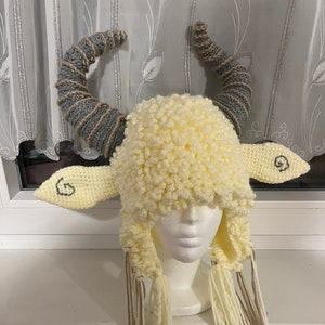 May include: A crocheted hat shaped like a sheep with gray horns and long white and brown tassels. The hat is on a white mannequin head.