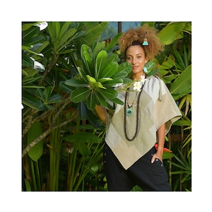 Könnte beinhalten: Eine Frau trägt einen beigefarbenen Poncho mit einer braunen Perlenkette und einem türkisfarbenen Anhänger. Sie steht vor einer üppigen grünen tropischen Pflanze.