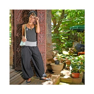 May include: A person wearing a gray tank top, black harem pants, and a light blue purse. The person is leaning against a carved wooden door. The background includes lush greenery and stone structures.