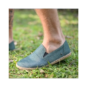 May include: A pair of dark green canvas slip-on shoes with a rope sole. The shoes are being worn by a person standing on grass.