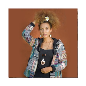 Könnte beinhalten: Eine Frau mit lockigem braunen Haar trägt eine bunte Patchworkjacke mit einem schwarzen Kapuzenpullover darunter. Sie trägt eine schwarze Halskette mit einem Anhänger und Ohrringe. Sie hat eine weiße Blume im Haar.