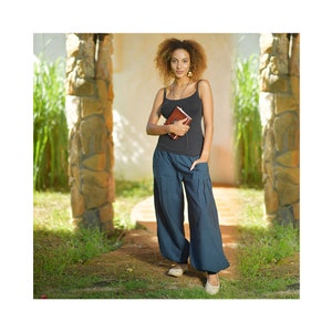 Puede incluir: Una mujer con un top negro sin mangas y pantalones azules anchos. Est&aacute; sosteniendo un peque&ntilde;o libro marr&oacute;n encuadernado en cuero en sus manos. Est&aacute; de pie en un entorno de jard&iacute;n con pilares de piedra y vegetaci&oacute;n verde.